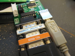 Photo of adapter board with a nine pin serial cable, null modem, gender changer, and USB cable attached.